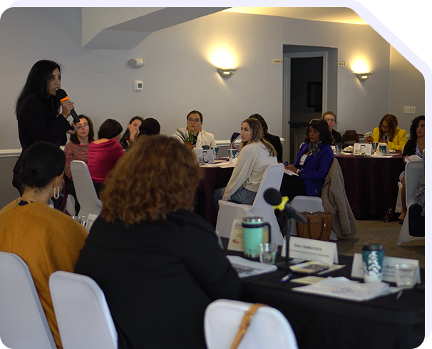 Professional business meeting or conference room photograph showing multiple participants seated around a dining table, with a woman standing and presenting to the group. The modern meeting space features warm wall lighting, a curved ceiling detail, and professional tableware. Used to represent Star Trak Investments LLC's client meetings, investor workshops, business conferences, team collaboration, and professional networking events for wealth management and financial advisory services