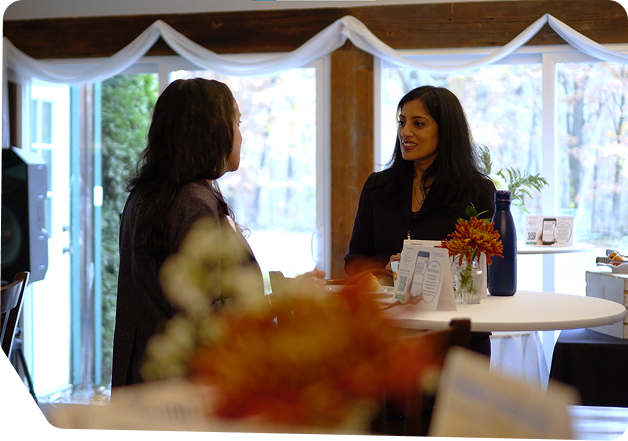 Professional one-on-one consultation or client meeting photograph showing two businesswomen conversing at a desk with a potted orange flower arrangement in the foreground. The bright, modern office space features elegant blue and white draped fabric, natural window lighting, and professional furnishings, representing Star Trak Investments LLC's personalized financial advisory sessions, wealth management consultations, and client relationship building for investment services