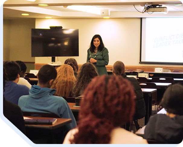 Educational seminar photograph showing a female instructor in a green blazer standing at the front of a classroom, presenting to a diverse audience of seated students and professionals. The classroom features fluorescent ceiling lights, a projection screen, and institutional furnishings, representing Star Trak Investments LLC's commitment to investor education, financial literacy workshops, professional development training, and client knowledge-building seminars on investment strategies and wealth management