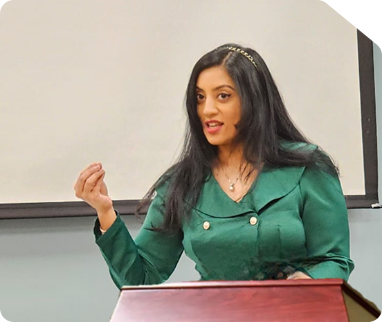 Professional photograph of a confident businesswoman wearing a green blazer and formal attire, speaking at a podium or presentation table with a microphone, against a neutral classroom or conference room backdrop. The image represents Star Trak Investments LLC's investment advisors, female financial leadership, educational seminars, and client engagement in wealth management and financial planning services